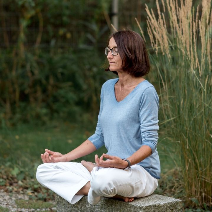 Praxisinhaberin in Meditation sitzend auf einem Stein im Garten